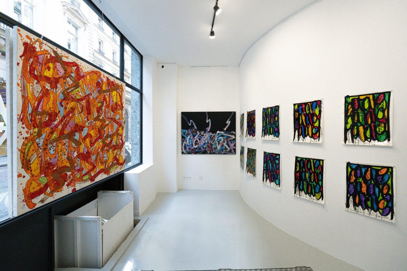  Perspective le long de la vitrine, avec une composition orange à gauche et de petits formats multicolores sur le mur de droite.