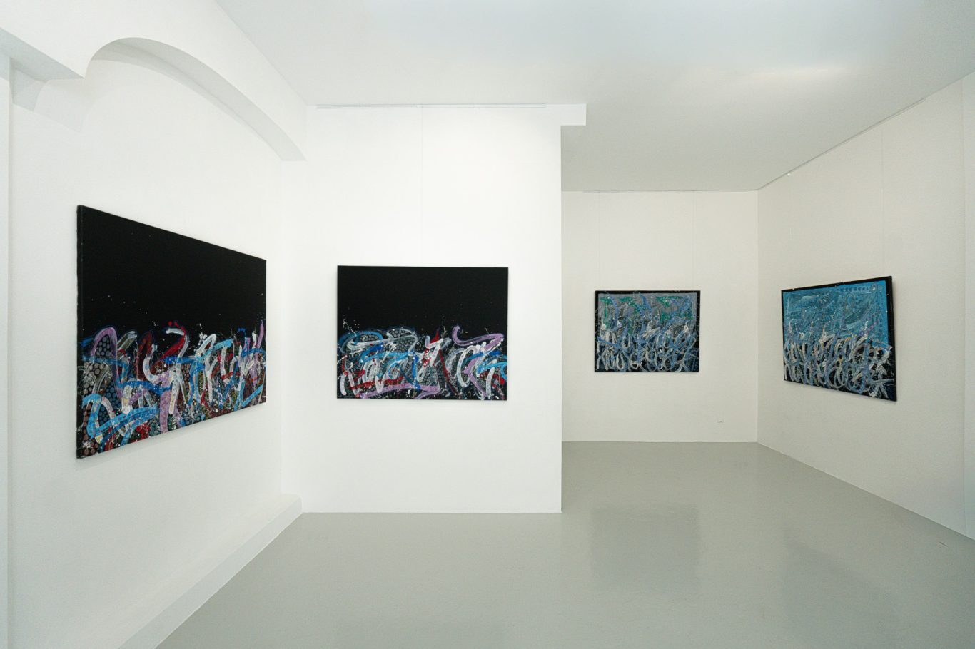 Vue d’un angle de la galerie avec des compositions sur fond noir et des œuvres à dominante bleue.