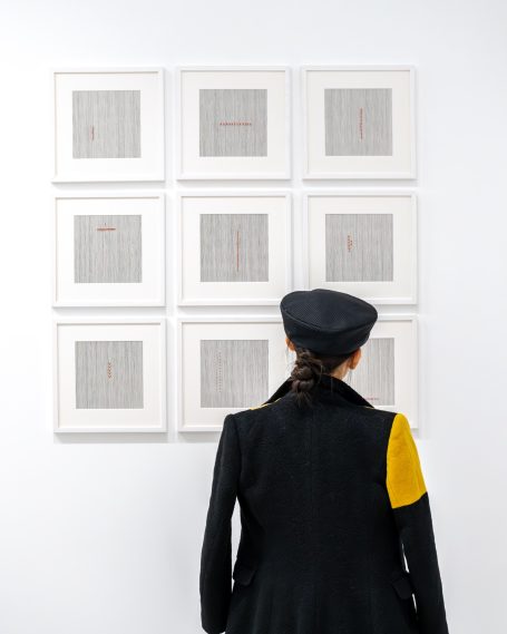 Une personne avec un chapeau noir regarde des œuvres encadrées sur un mur blanc.