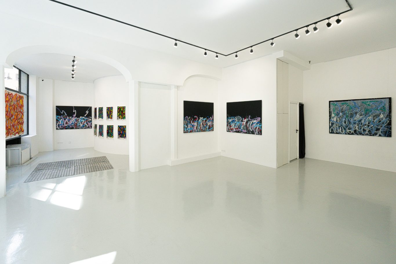 Vue d’ensemble de la galerie aux murs blancs, avec une série de petits formats à gauche et deux œuvres sur fond noir au centre.