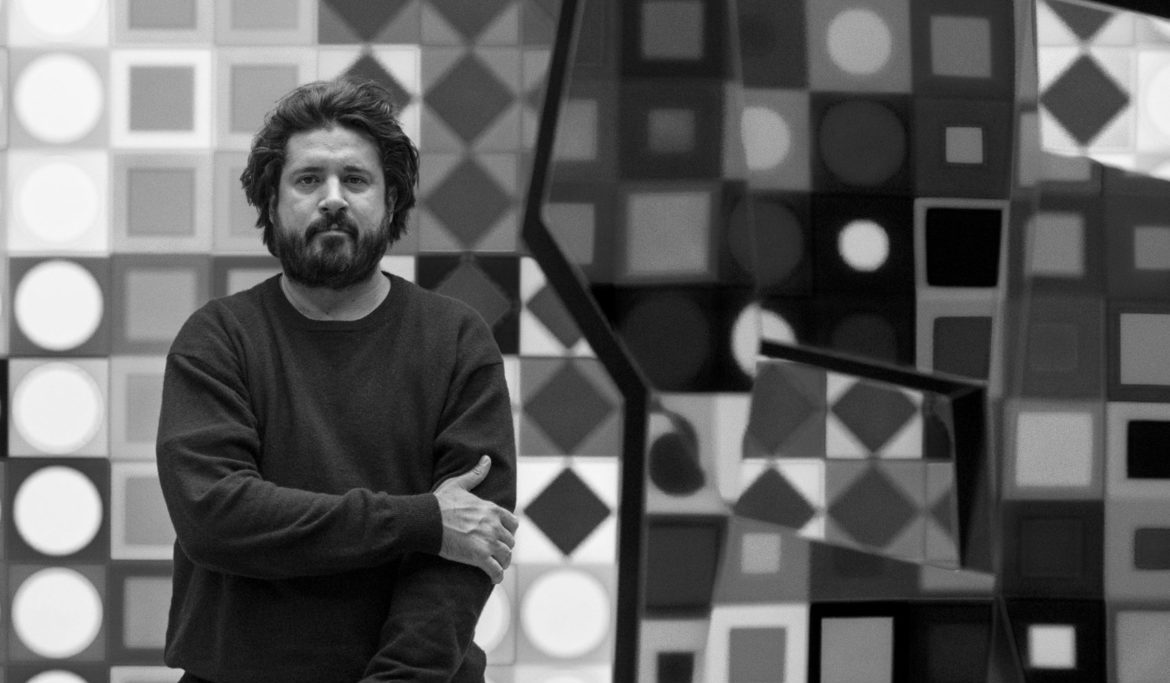 Portrait en noir et blanc de l’artiste Arthur Dorval devant ses œuvres géométriques présentées lors de l’exposition Fata Morgana chez H.D Ga